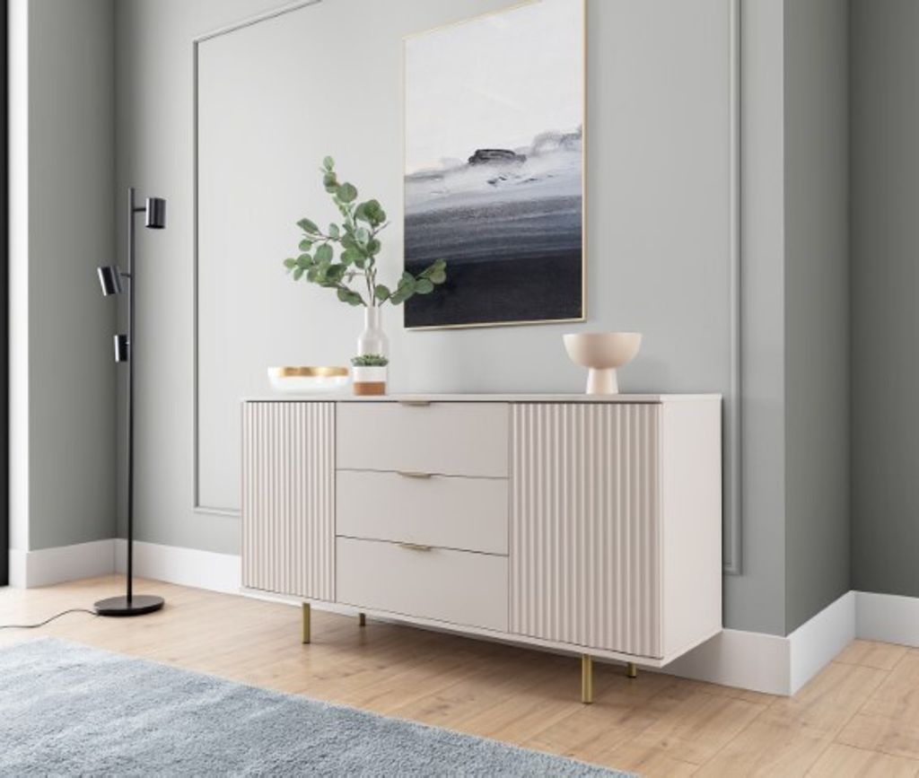 A modern sideboard with fluted doors and drawers, set against a light gray wall with a framed abstract painting above it. A floor lamp stands to the left, and a blue rug is visible in the foreground.