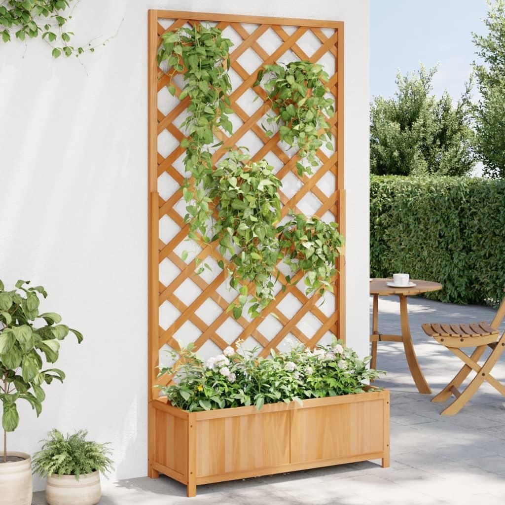 A wooden planter box with a trellis attached, filled with plants, stands against a white wall on a patio. A small round table and chair are visible in the background.
