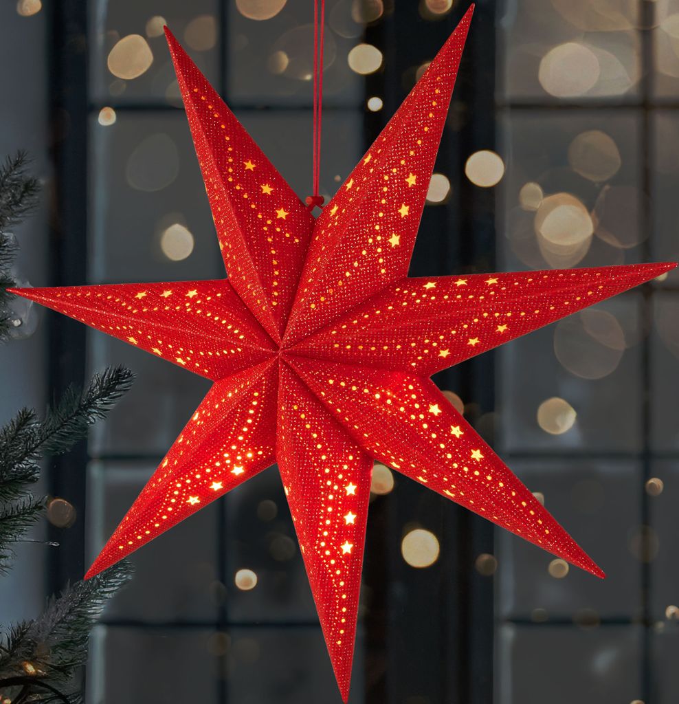 A red 3D star-shaped Christmas lantern with small yellow stars and dots, illuminated from within, hangs in front of a window with blurred lights.