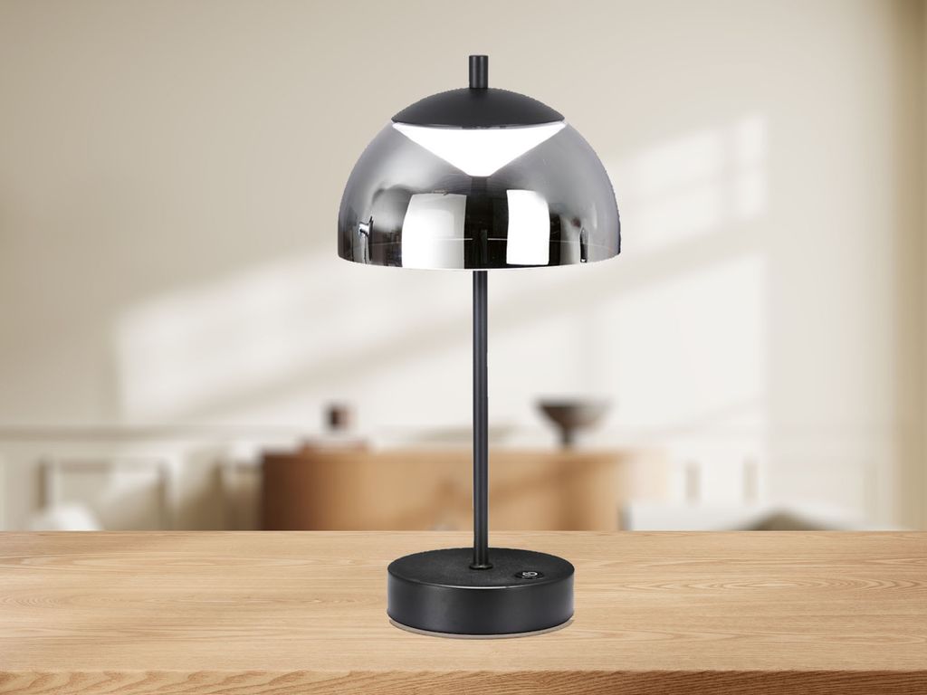 A modern black table lamp with a reflective silver dome shade is placed on a wooden table in front of a blurred interior background.