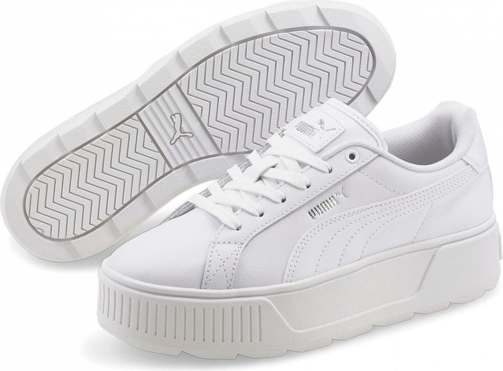 A pair of white Puma Karmen L women's sneakers are shown from a slightly elevated angle, with one shoe in the foreground and the sole of the other shoe visible behind it.