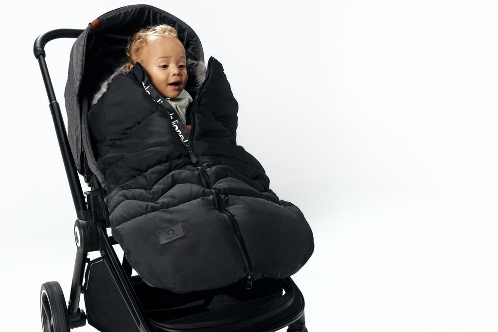 A baby wrapped in a black footmuff is sitting in a stroller against a white background.