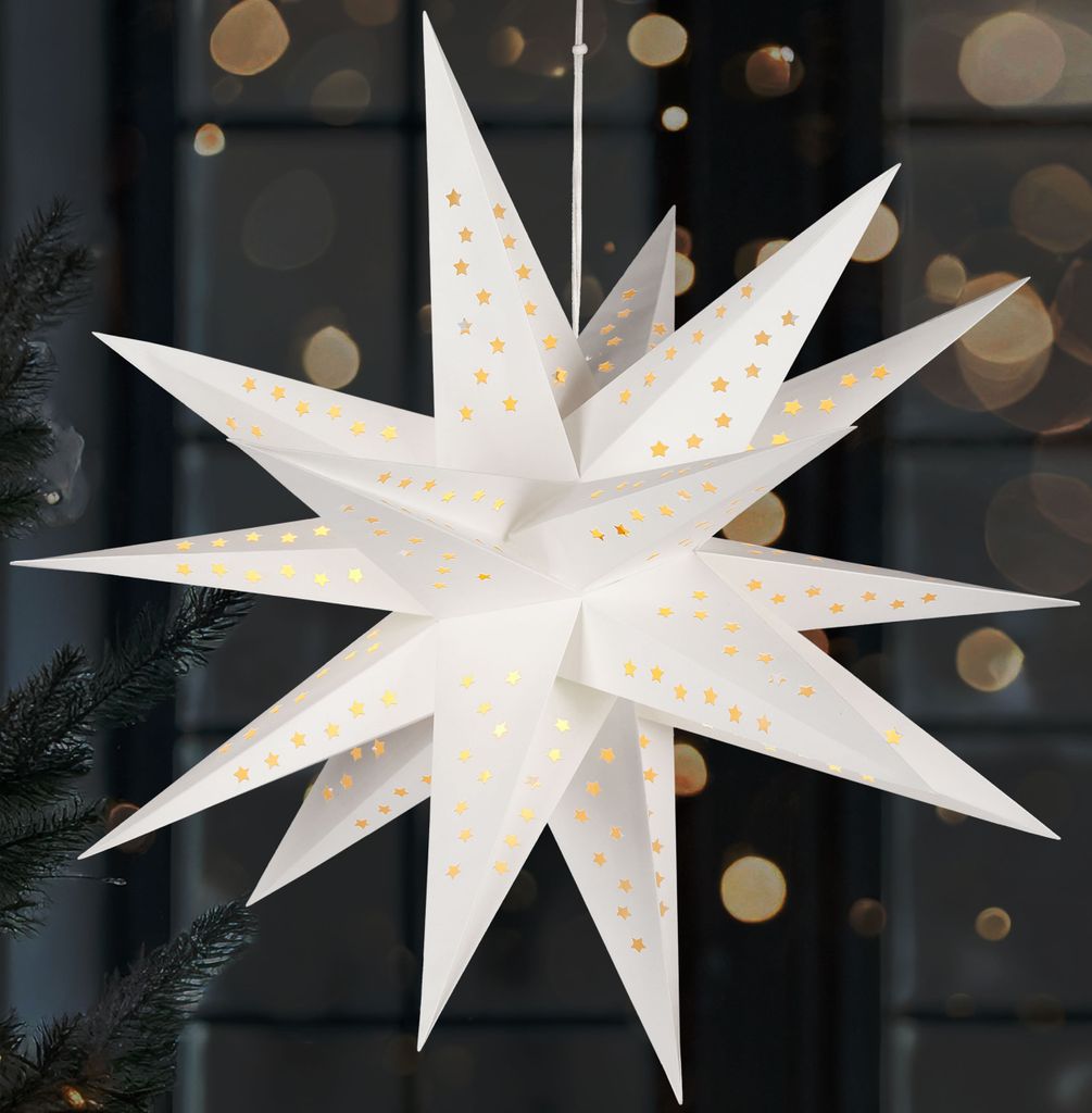 A white paper star lantern with gold star cutouts hangs in front of a dark background with bokeh lights.