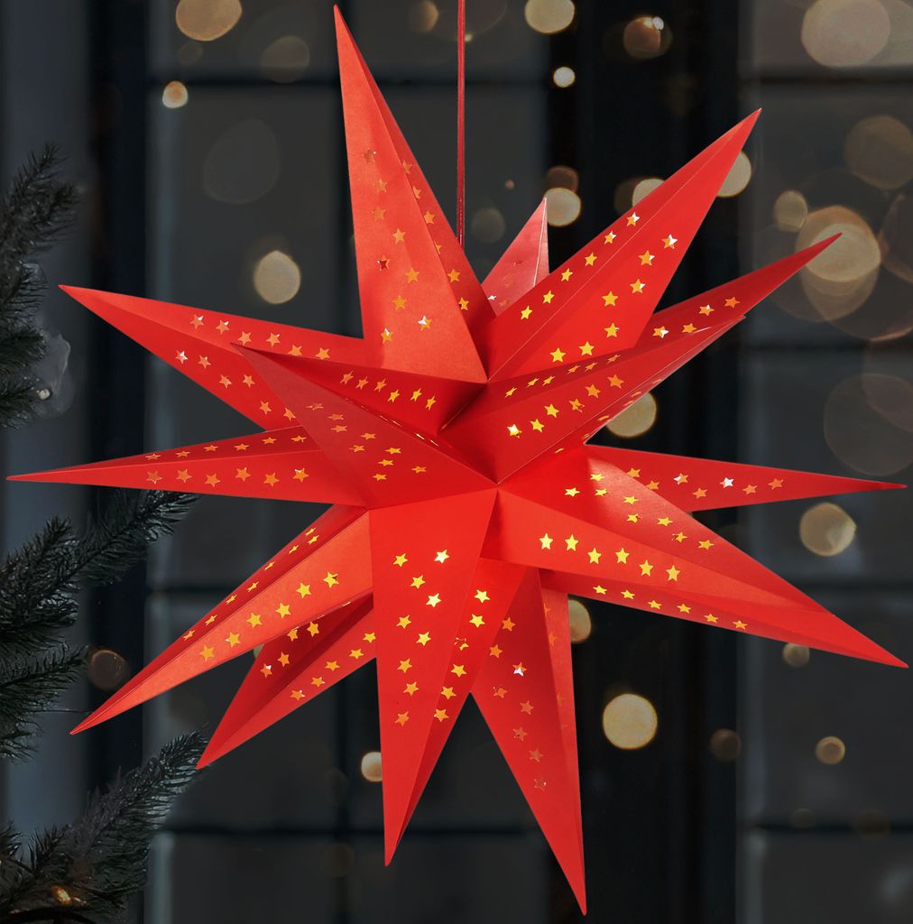 A red, star-shaped paper lantern with small gold stars cut out of it, hanging in front of a blurred background with bokeh lights.