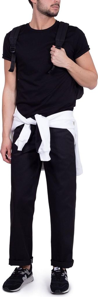 A man wearing black Dickies work pants and a black t-shirt, with a white sweatshirt tied around his waist and a black backpack.