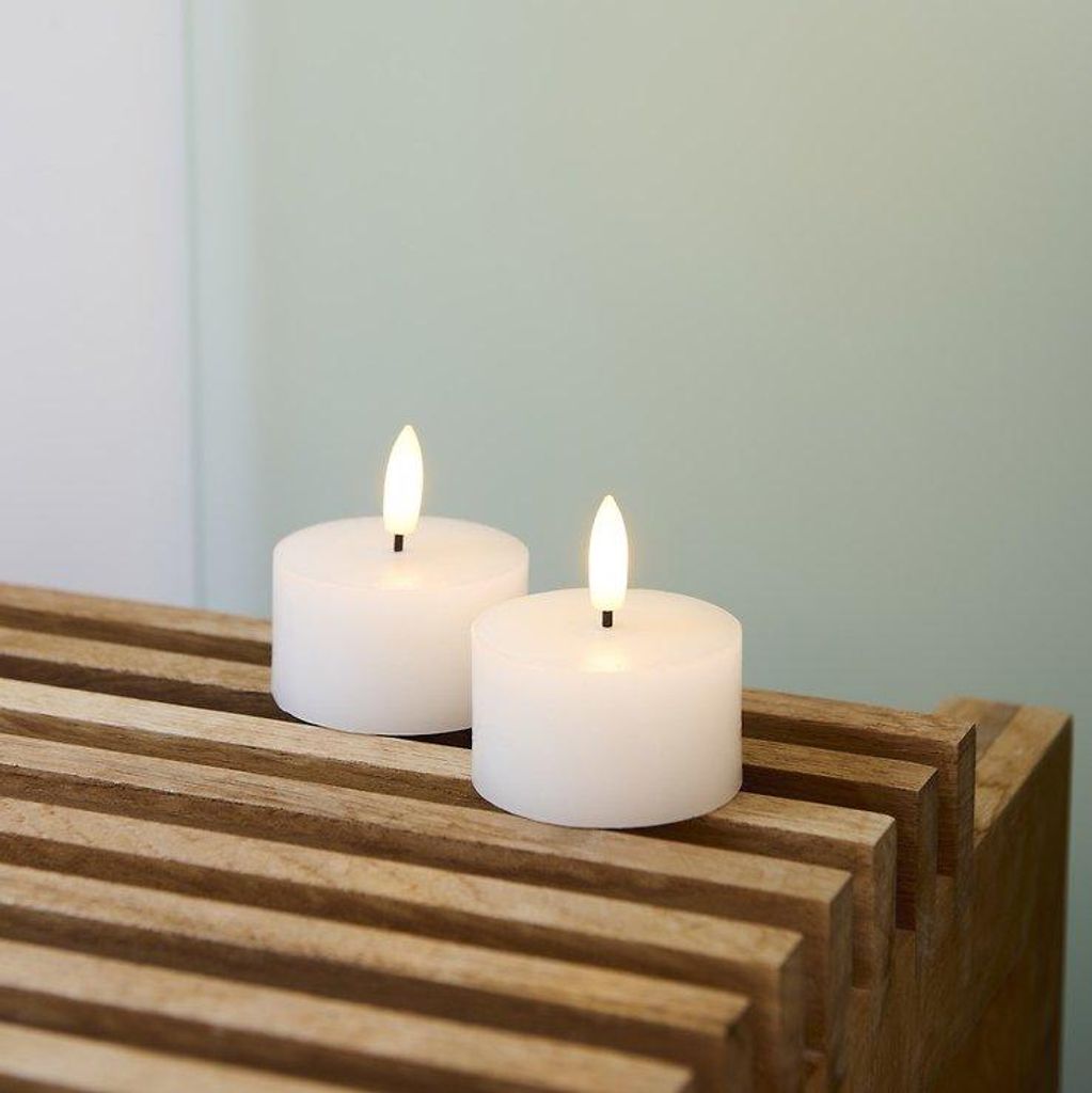 Two white LED tea lights with illuminated flames sit on a slatted wooden surface against a pale green and white background.