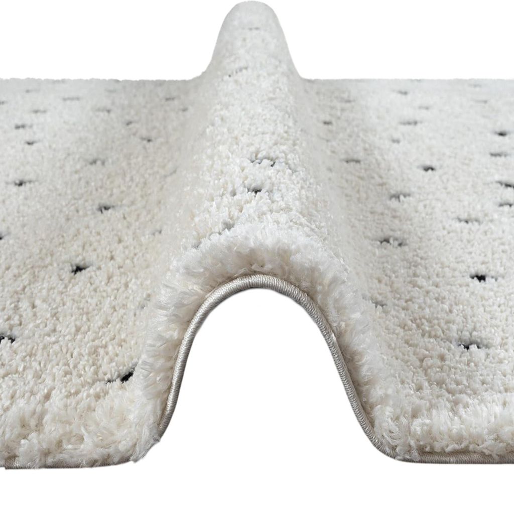A close-up, angled view of a beige rug with black dots, showing its high pile texture and a rolled edge.