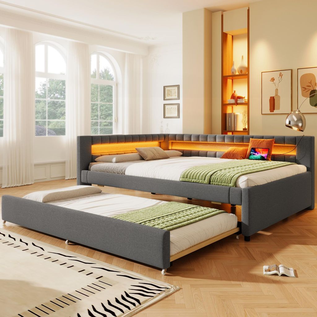 A bedroom scene featuring a grey upholstered daybed with an integrated LED light strip and a pull-out trundle bed. The room has large arched windows with sheer curtains and a wooden shelf unit with decorative items.