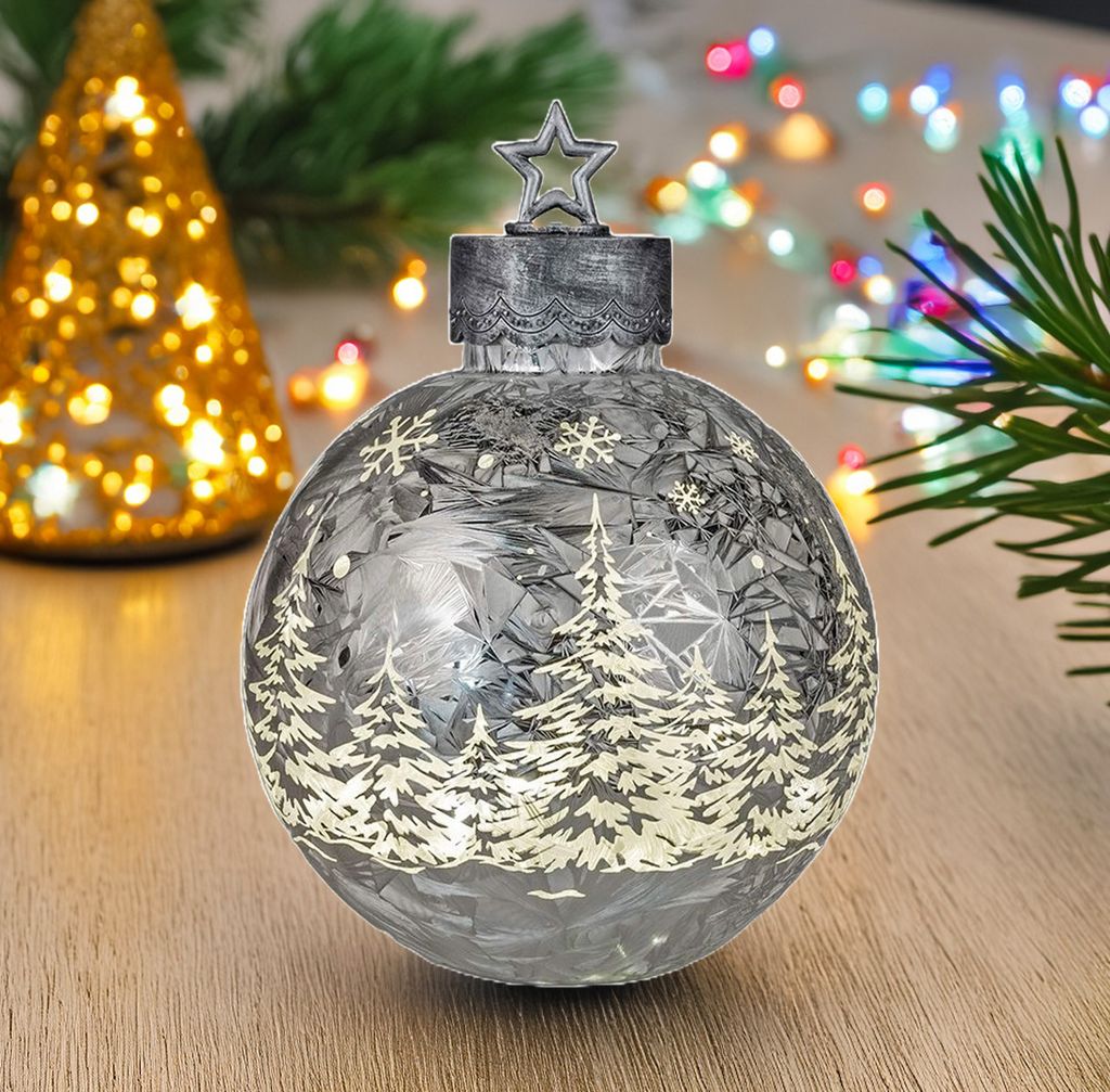 A decorative Christmas ornament with a winter forest scene and snowflakes, lit by internal LEDs, sits on a wooden surface. In the background, a blurred golden Christmas tree with lights and colorful string lights are visible.