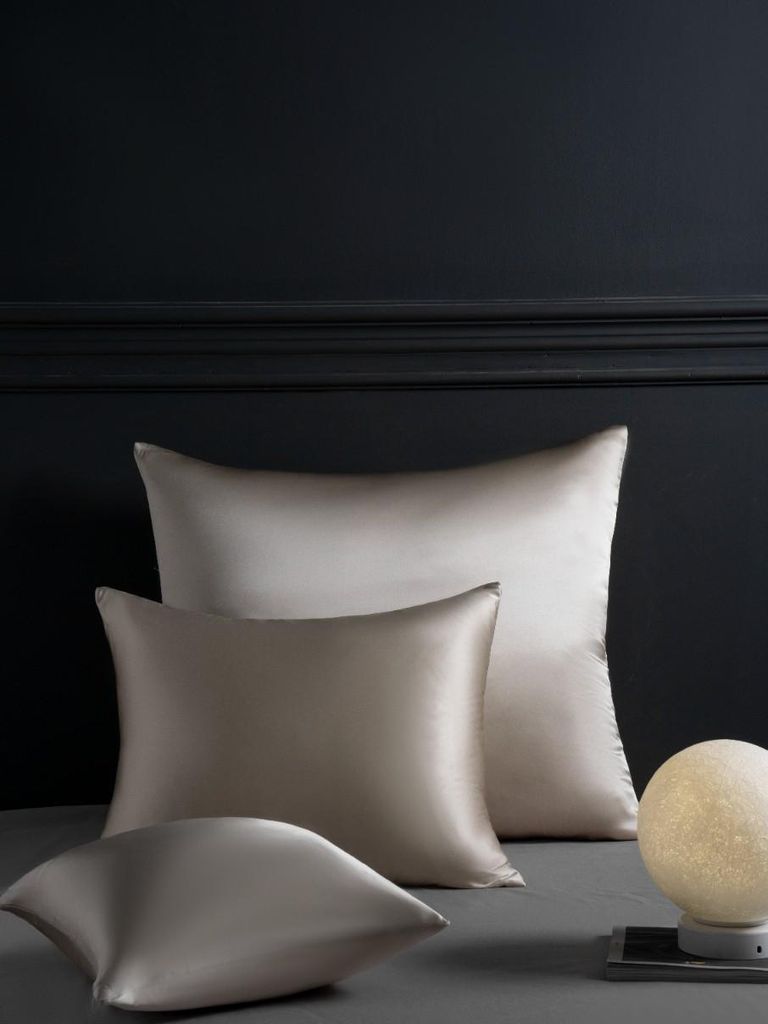 Three silk pillowcases are arranged on a surface in front of a dark wall with decorative molding. A small, round lamp is visible on the right.