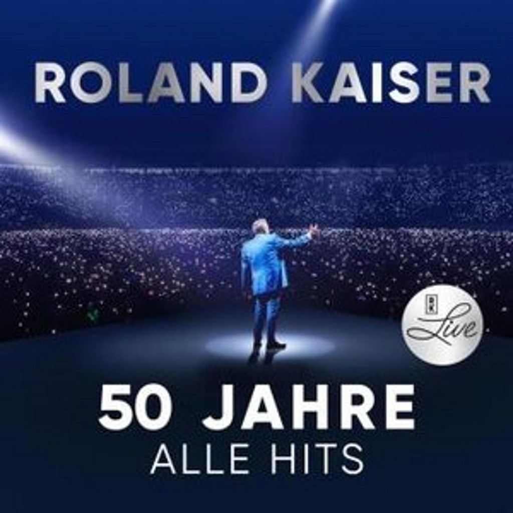 A man in a blue suit stands on a stage in front of a large audience, illuminated by spotlights. Text indicates it's a live album celebrating 50 years of hits.