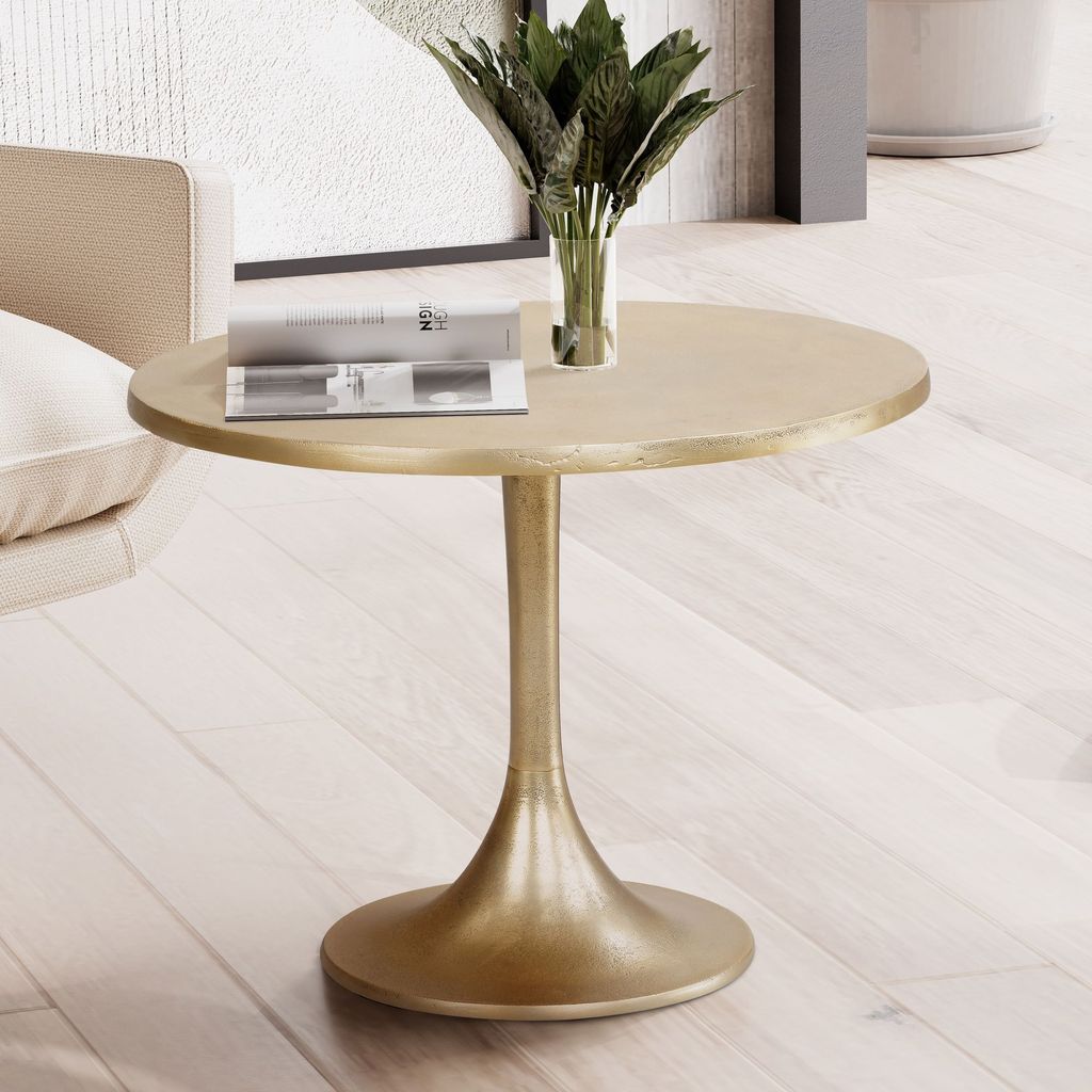 A gold tulip-style side table with a vase of greenery and a magazine on top, placed on a light wood floor next to a chair.