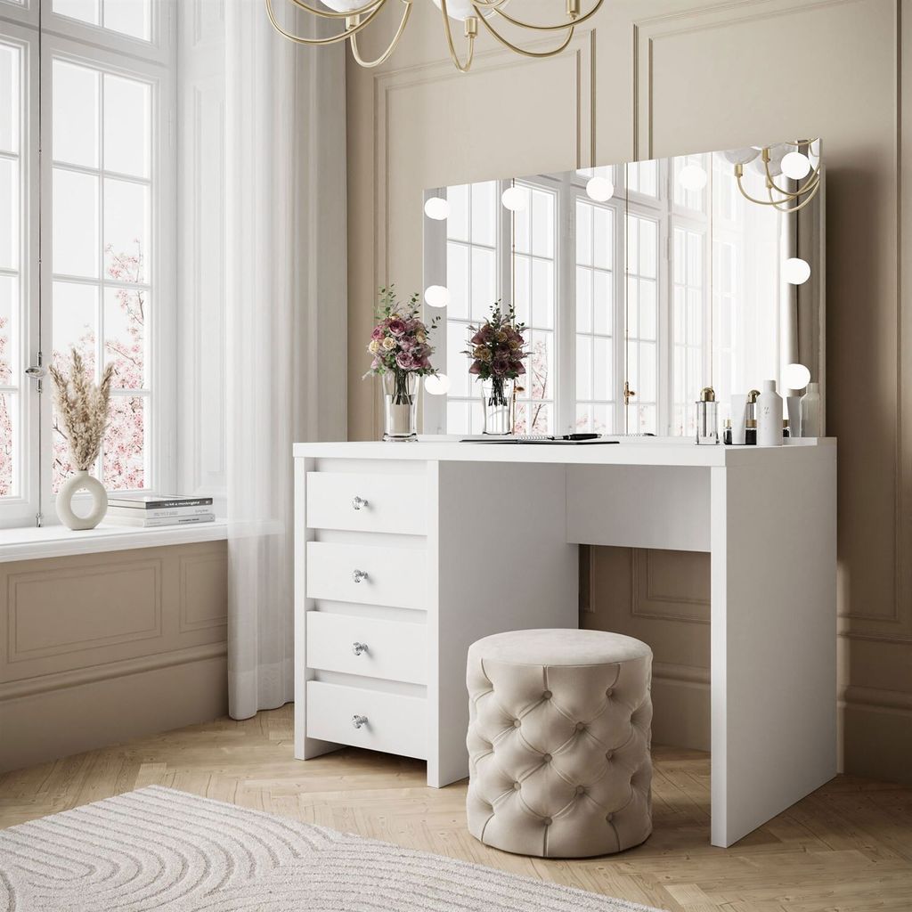 A white vanity table with a mirror and lights, a tufted stool, and decorative flowers in vases, set in a well-lit room with a large window.