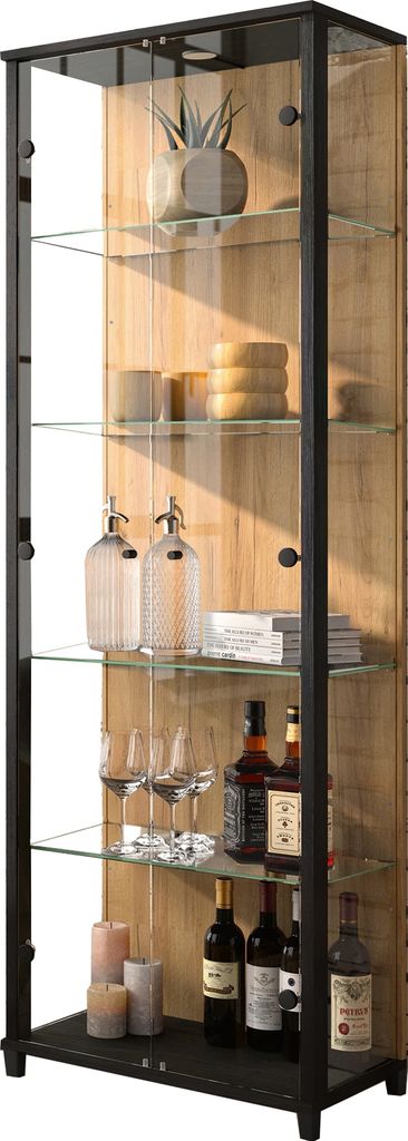 A tall, narrow glass display cabinet with a black frame and wood-effect back panel, filled with various items like bottles, glasses, and decorative objects.