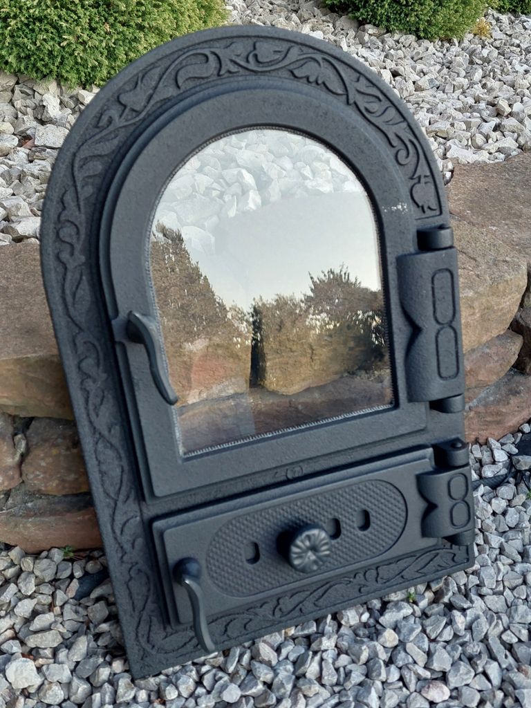 A black cast iron oven door with a glass window, placed outdoors on gravel and rocks.