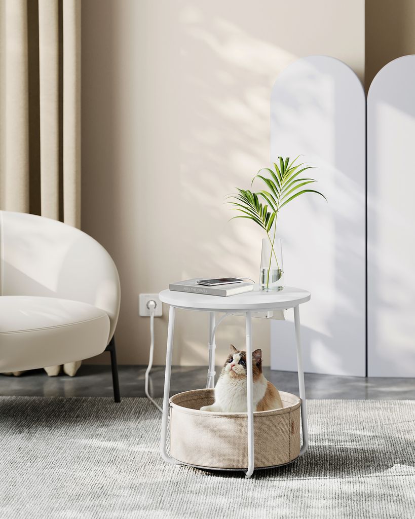 A cat sitting in a round pet bed integrated into a round side table, with a chair and curtains in the background.