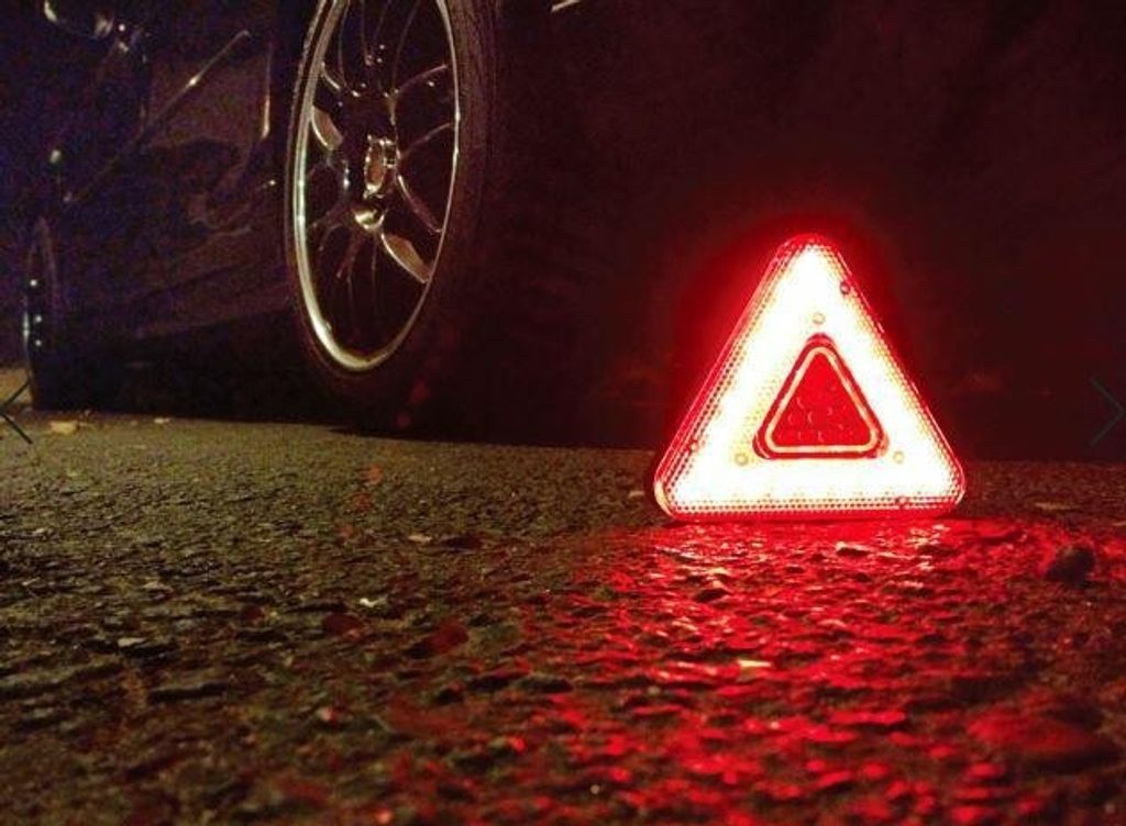 A red warning triangle is placed on the asphalt next to a car tire at night, indicating a breakdown.