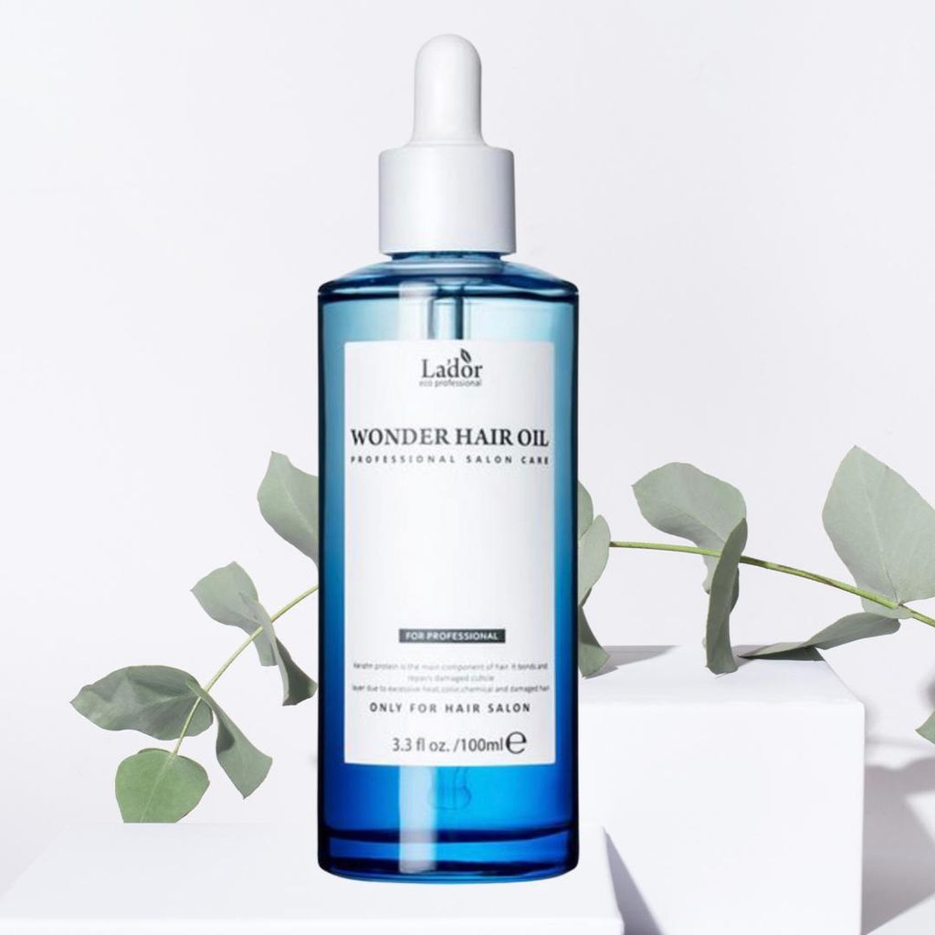 A bottle of La'dor Wonder Hair Oil is displayed on a white pedestal, with green leaves in the background.