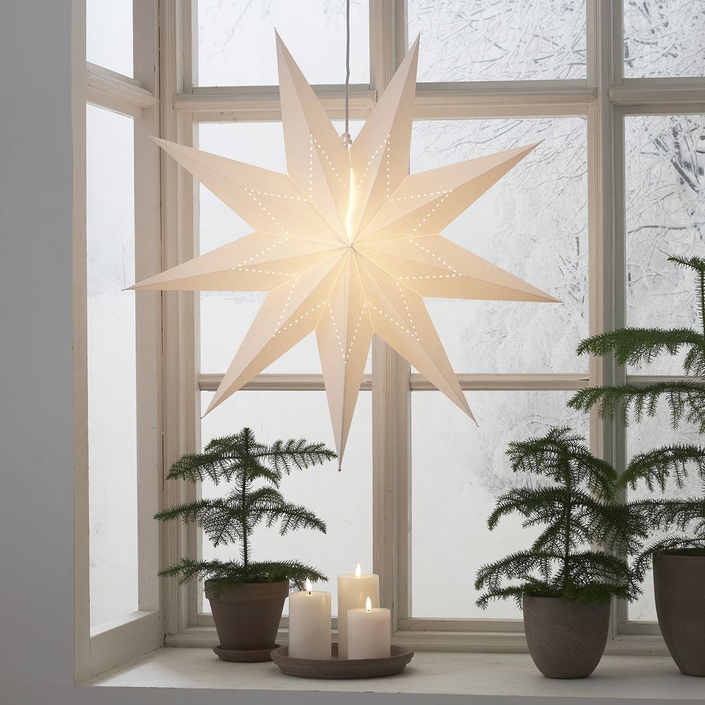A paper star lantern hangs in a window with small Christmas trees and candles on the windowsill.