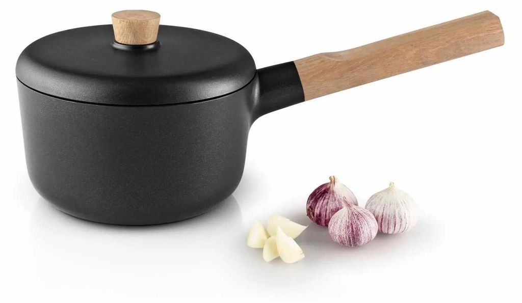 A black saucepan with a wooden handle and lid sits on a white surface next to three heads of garlic and some peeled garlic cloves.