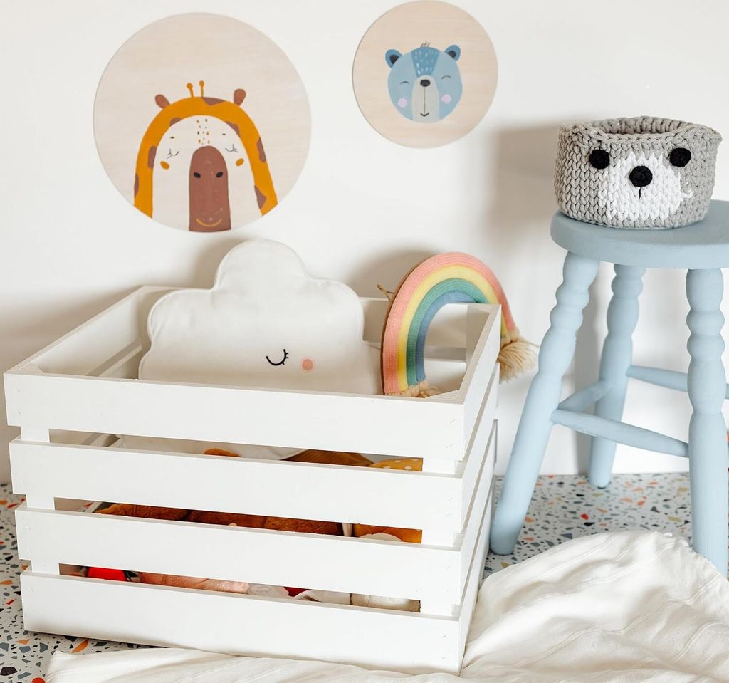 A white wooden crate filled with children's toys, including a cloud pillow and a rainbow decoration, is placed on the floor next to a light blue stool with a knitted basket on top. Two circular wooden wall decorations with animal illustrations are visible 