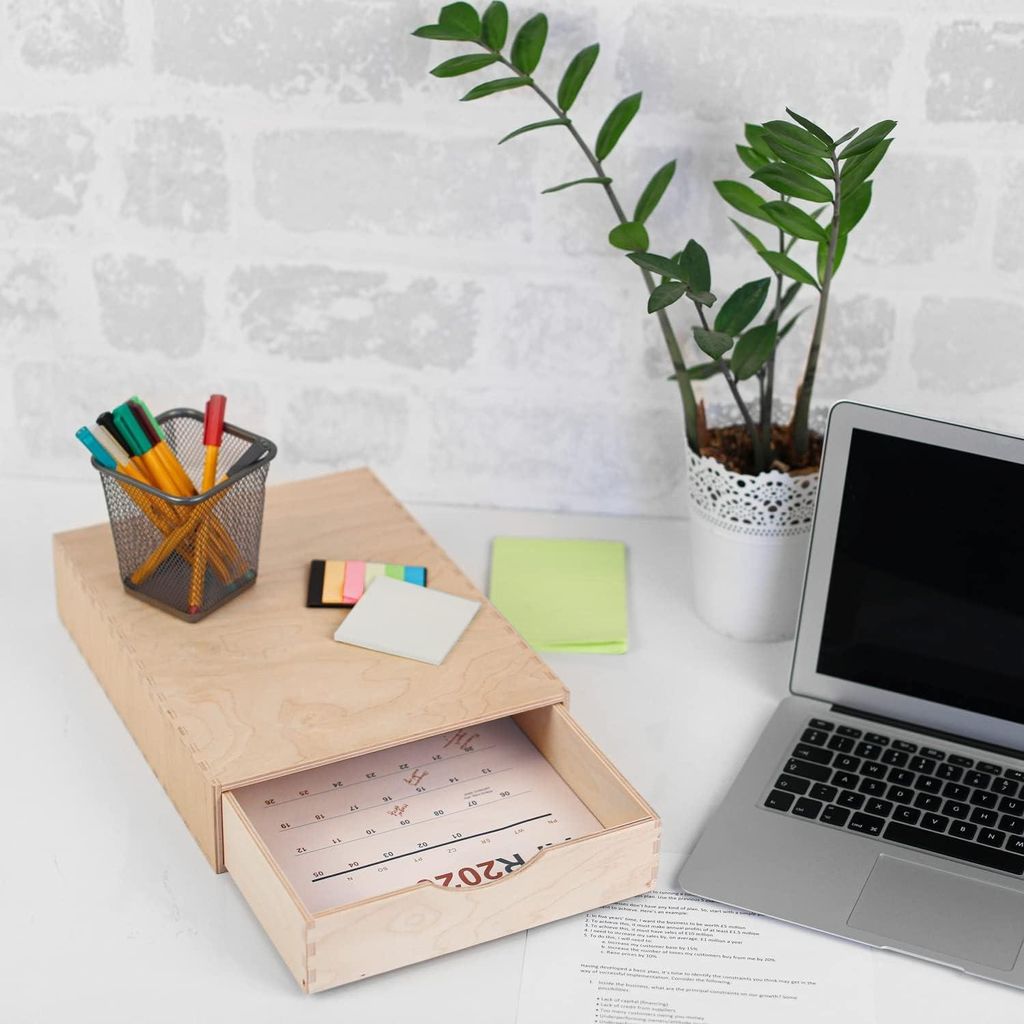A wooden desk organizer with an open drawer containing a calendar, placed on a white surface with a laptop, a potted plant, and some sticky notes in the background.