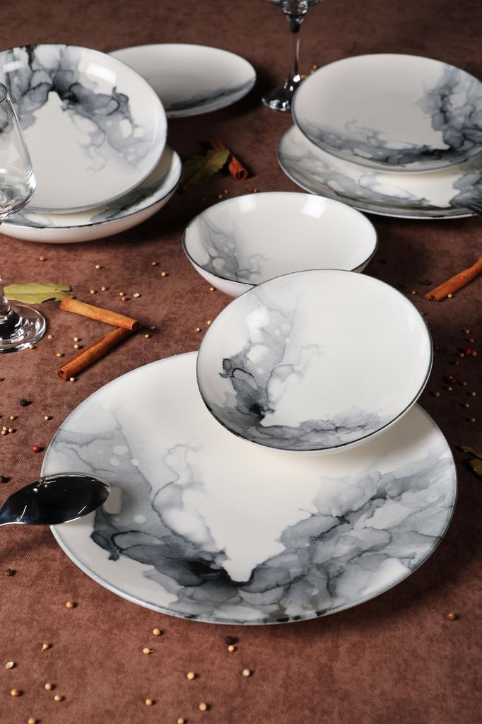 A collection of ceramic plates and bowls with a marbled gray and black design, arranged on a brown surface with cinnamon sticks and other decorative elements.