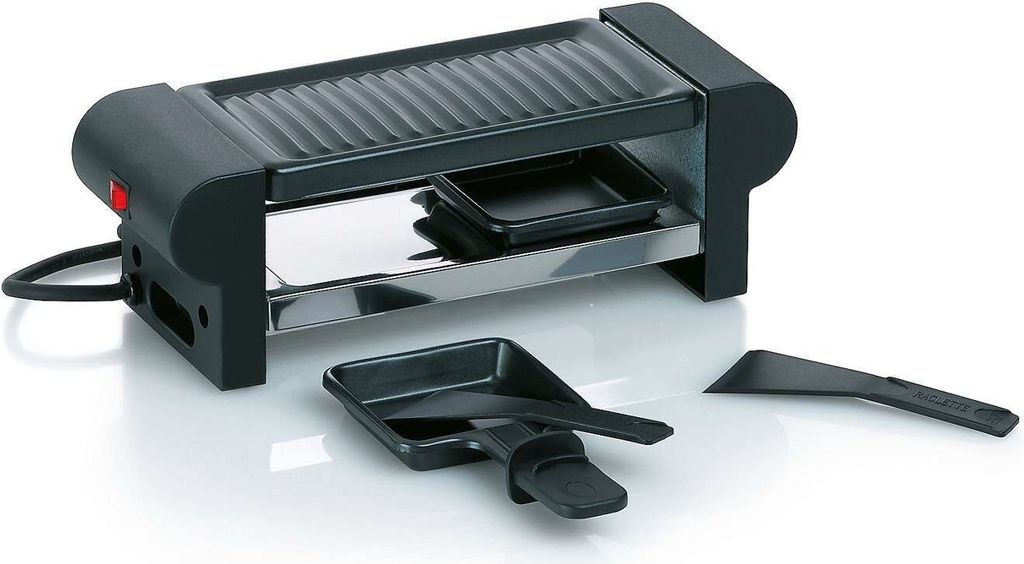 A black and silver electric raclette grill with two small black trays and a black spatula in front of it, set against a white background.