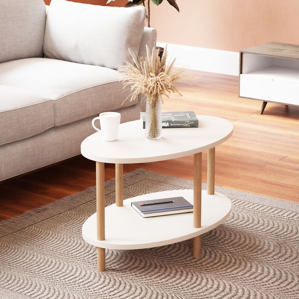 A white oval coffee table with wooden legs is placed on a patterned rug in a living room setting, next to a sofa. A vase with dried flowers and a mug are on the table.