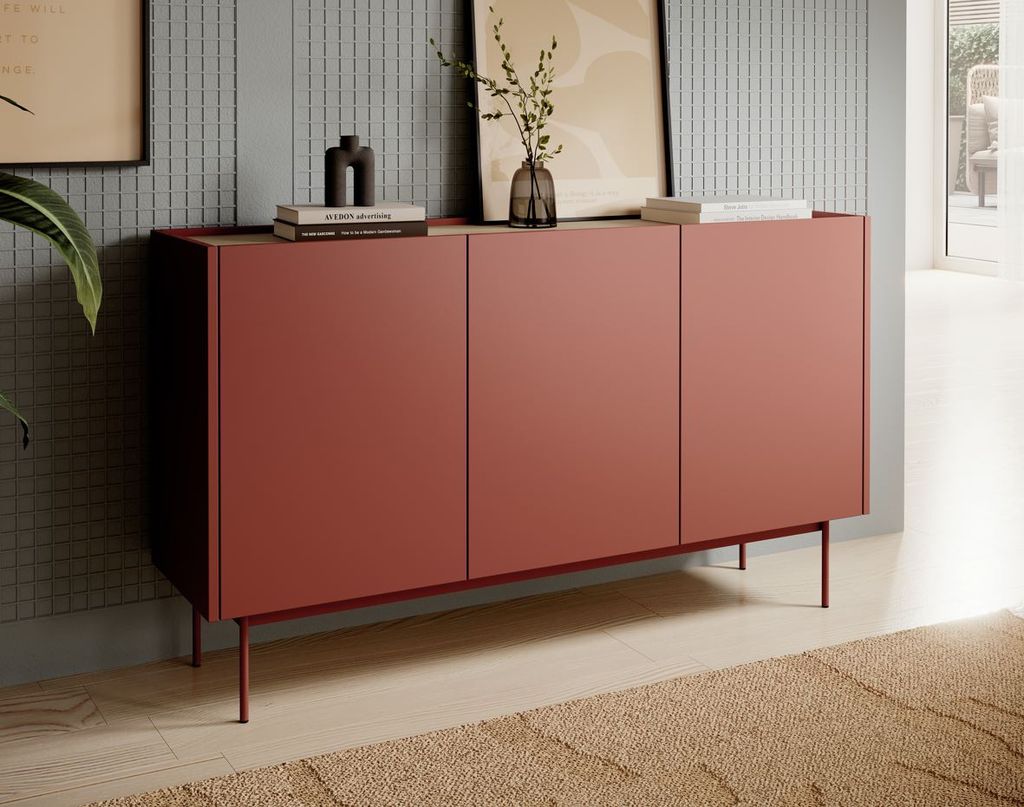 A modern red sideboard with three doors stands against a textured gray wall, adorned with framed art and decorative items on its surface. The scene is set in a well-lit room with a beige rug in the foreground.