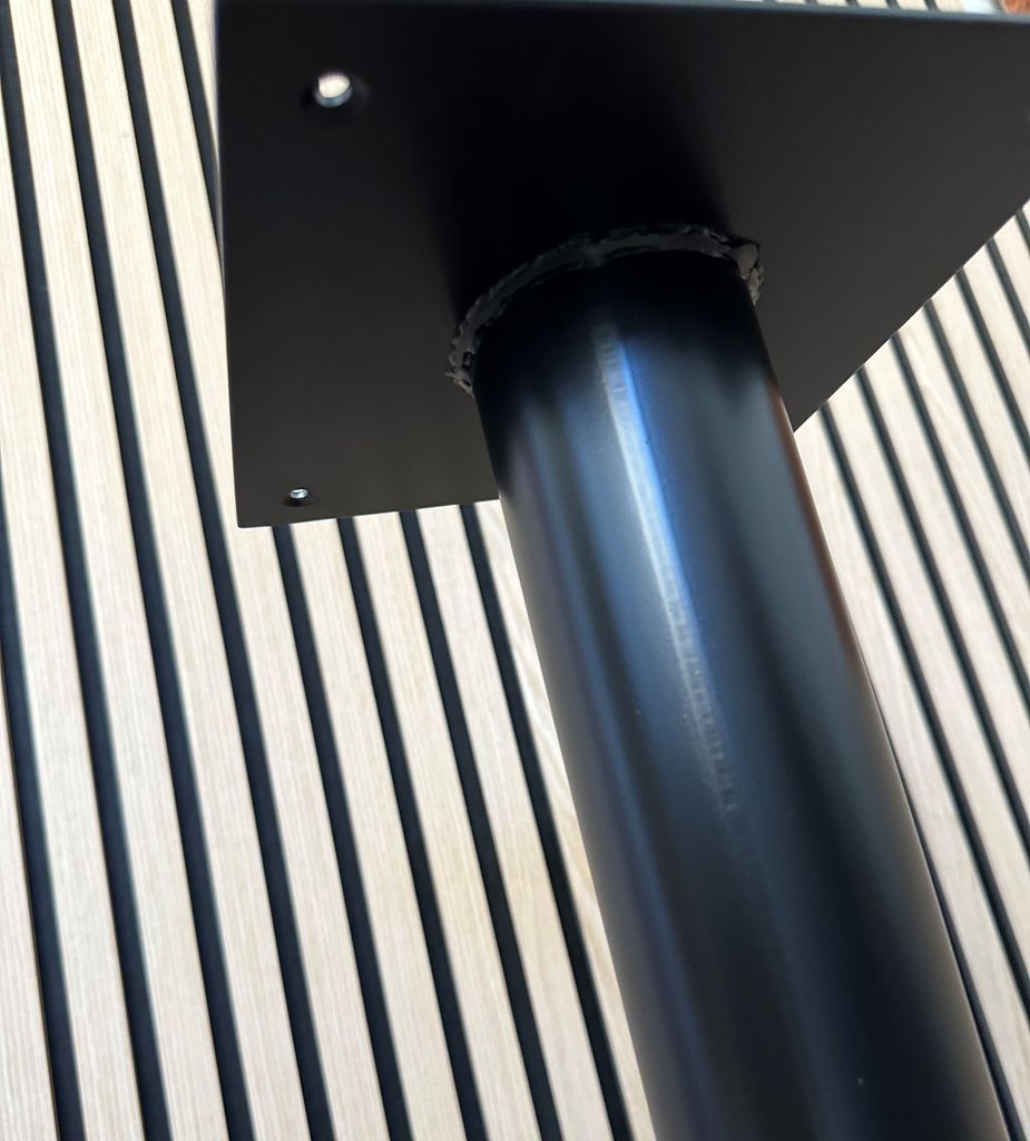 A close-up of a black metal table base with a square mounting plate, set against a background of light-colored wooden slats with dark vertical stripes.