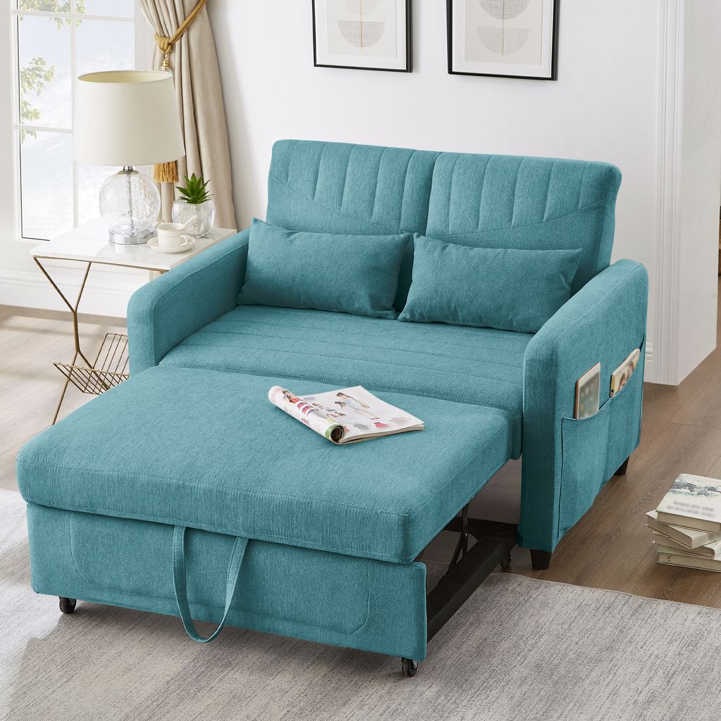 A teal sofa bed is shown in a living room setting, with a side table and lamp to the left, and books to the right.