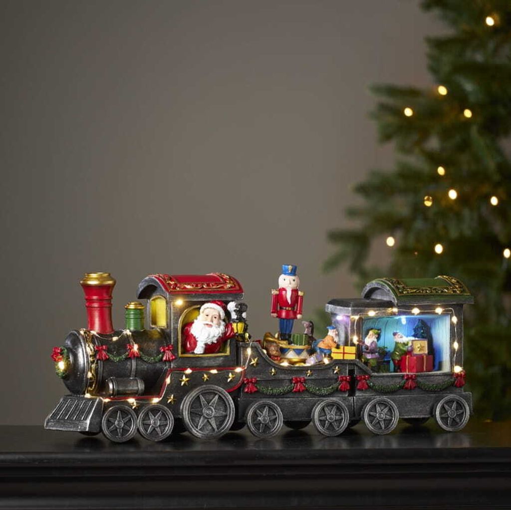 A festive Christmas train decoration with Santa Claus and nutcracker figures, illuminated by small LED lights, placed on a dark surface with a blurred Christmas tree in the background.