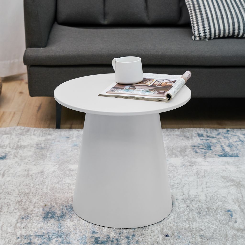 A white round side table with a magazine and a mug on it, placed on a rug in front of a sofa.