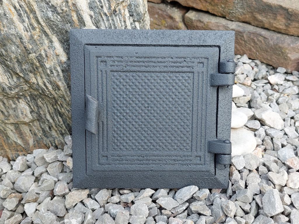 A cast iron access door for a chimney or oven, placed on a bed of gravel with rocks in the background.