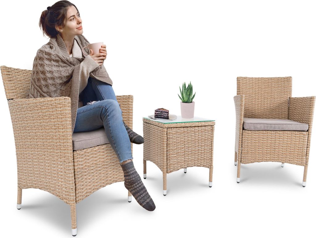 A woman sits in a wicker chair, wrapped in a blanket and holding a mug. A small table with a cake and a plant sits next to her, with another wicker chair to the right.