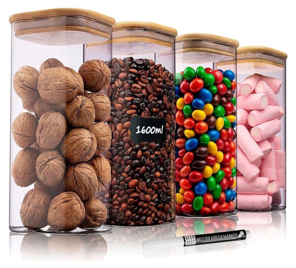 Vier klare Glasvorratsbehälter mit quadratischen Bambusdeckeln, gefüllt mit Walnüssen, Kaffeebohnen, bunten Bonbons und rosa Marshmallows, mit einem weißen Kreidestift davor