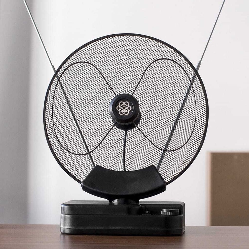 A black indoor TV antenna with a circular mesh screen and two extendable rods is placed on a wooden table.
