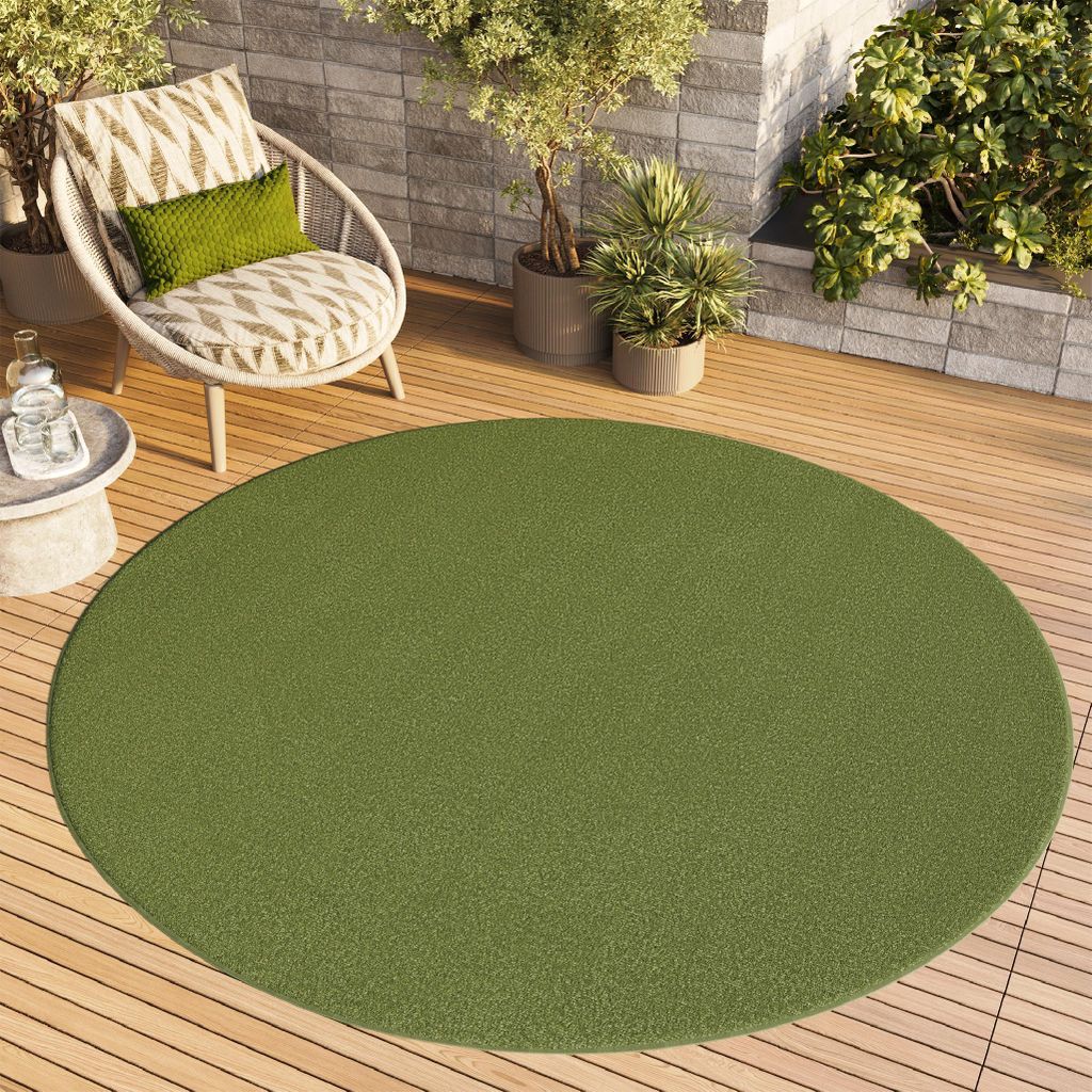 A round green rug on a wooden deck with a chair and plants.