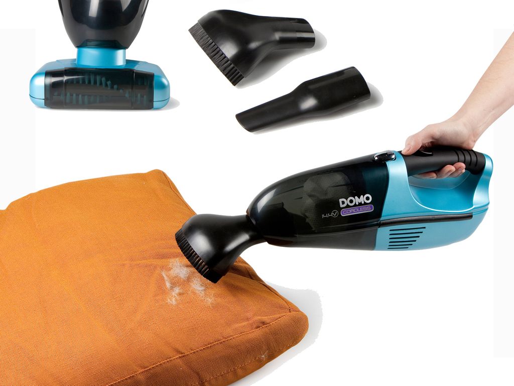 A hand holding a handheld vacuum cleaner with various attachments, demonstrating its use on an orange cushion.