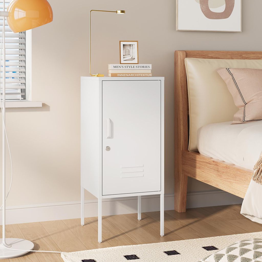 A white metal nightstand with a single door and a lock is placed next to a wooden bed frame. A floor lamp with an orange shade is to the left of the nightstand.