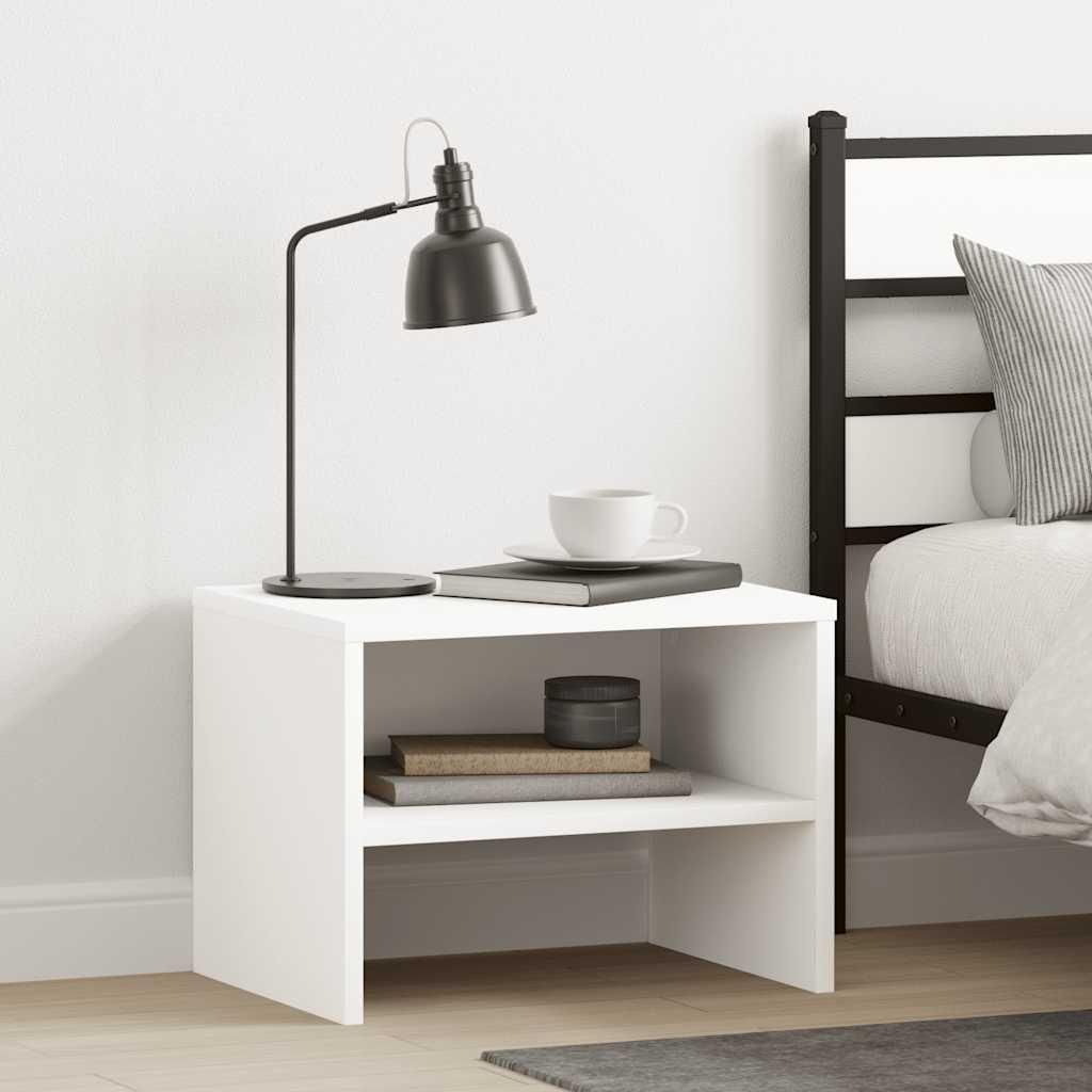 A white bedside table with a lamp, books, and a cup on it, next to a bed.