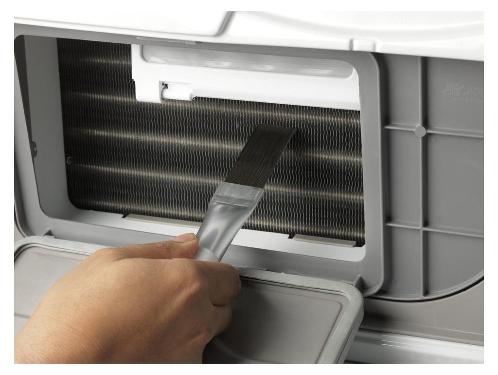 A hand is using a brush to clean the fins of an air conditioner.