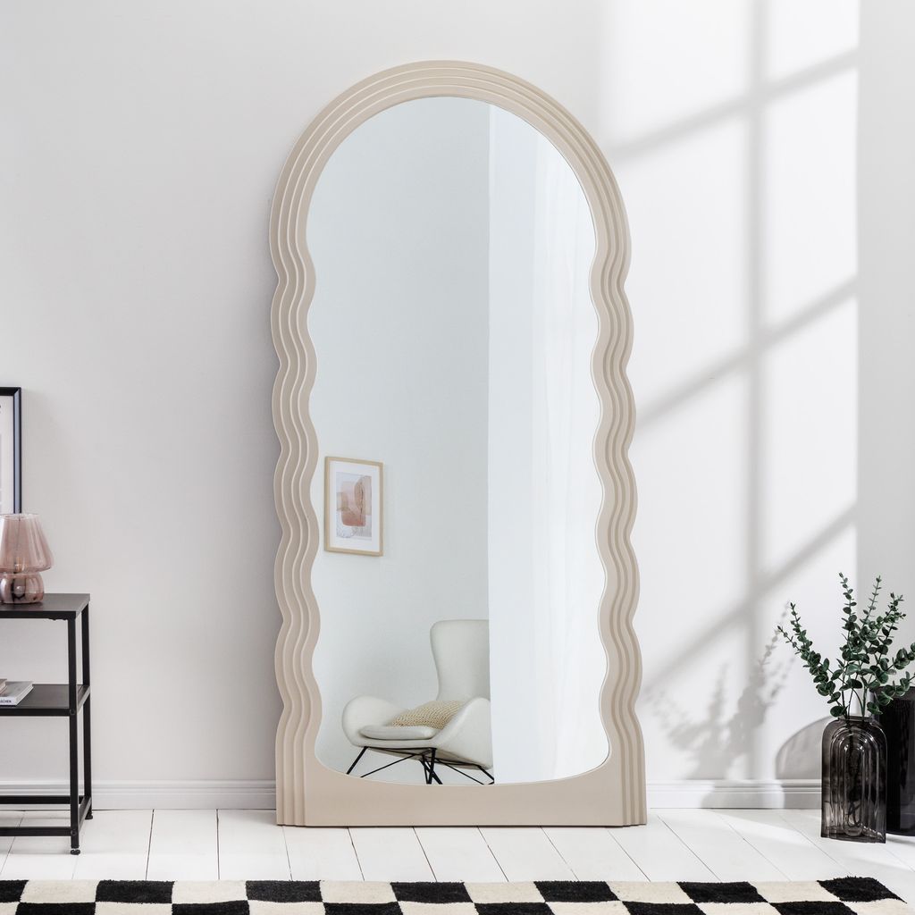 A full-length mirror with a wavy frame stands against a white wall, reflecting a chair and a framed picture. A small table with a lamp is to the left, and a checkered rug is on the floor.
