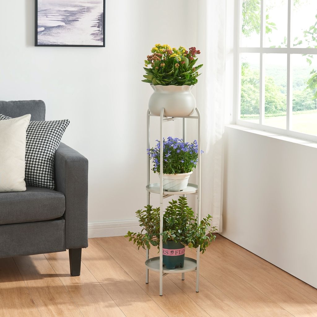 A plant stand with three potted plants is placed next to a gray sofa in a living room, with a window in the background.