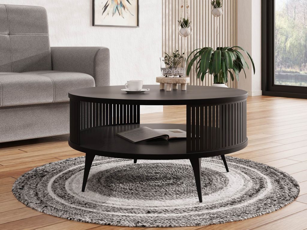 A black round coffee table with a slatted base and a lower shelf is placed on a round rug in a living room setting. A grey sofa is visible to the left, and a large window with greenery is in the background.