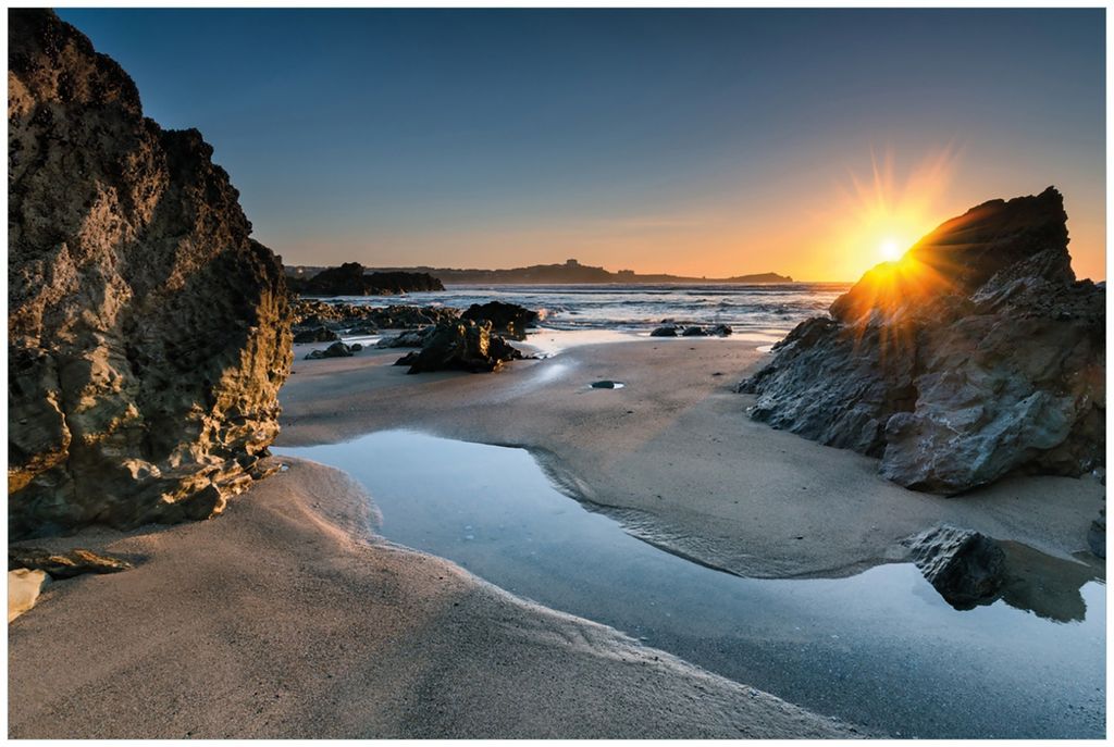 Wallario Premium Glasbild, freischwebende Optik, kräftige Farben, Größe 90 x 60 cm Motiv Sonnenuntergang hinter einem Felsen am Strand