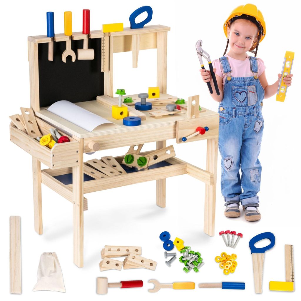 A young girl in overalls and a yellow hard hat stands next to a wooden toy workbench, holding tools.
