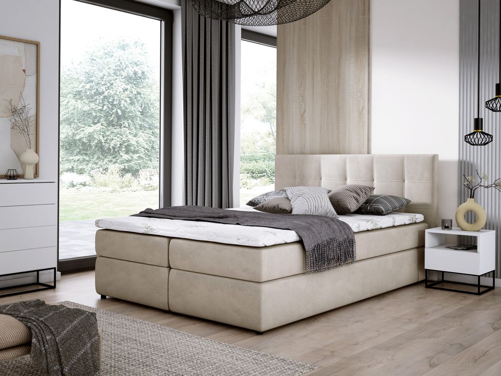 A bedroom scene featuring a beige boxspring bed with a mattress and pillows, set against a large window with a view of trees and a modern interior.