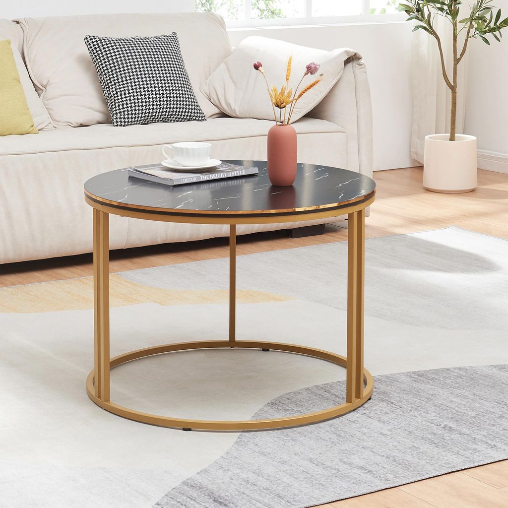 A round coffee table with a marble top and gold legs is placed on a rug in front of a sofa. A vase with dried flowers and a cup of coffee are on the table.